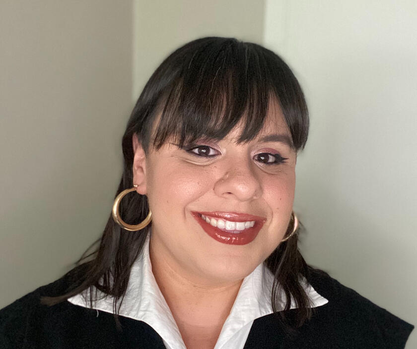 A headshot of Alejandra Martinez. She has medium length brown hair with bangs, hoop earrings, and a white collar peeking out of her sweater vest.